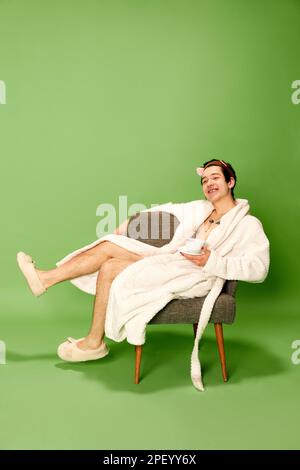 Portrait d'un jeune homme reposant portant un peignoir blanc et assis sur une chaise avec une tasse de café sur fond vert. Concept d'émotions positives Banque D'Images