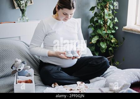 Femme enceinte avec grand ventre tenant body et triant des vêtements pour l'avenir. Comment organiser les vêtements pour nouveau-nés. Préparer les choses pendant la grossesse Banque D'Images