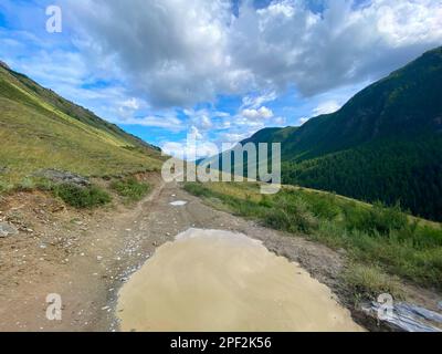 La route des voyageurs avec une grande flaque sur le bord de la colline près de la falaise va derrière la montagne sous les nuages puffy dans l'Altai à Siber Banque D'Images