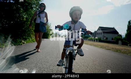 Mère enseigner à son fils à faire du vélo à l'extérieur sur une route urbaine. Garçon réussi portant un casque d'équilibrage et de pédaler par lui-même sur un vélo. Han Banque D'Images