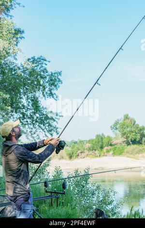 Pêcheur avec barbe et chapeau sur la rive de la rivière avec tige dans la main tirant une carpe Banque D'Images