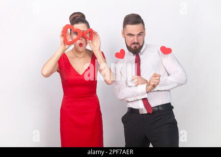 Portrait d'homme fort confiant en chemise blanche et femme surprise en robe rouge debout ensemble, tenant des figures de coeur, étant émotive. Prise de vue en studio isolée sur fond gris. Banque D'Images