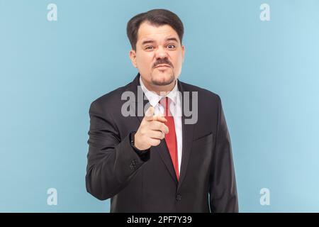 Portrait d'un triste homme bouleversé avec une moustache debout vous indiquant, regardant l'appareil photo avec visage frogné, portant un costume noir avec une cravate rouge. Prise de vue en studio isolée sur fond bleu clair. Banque D'Images