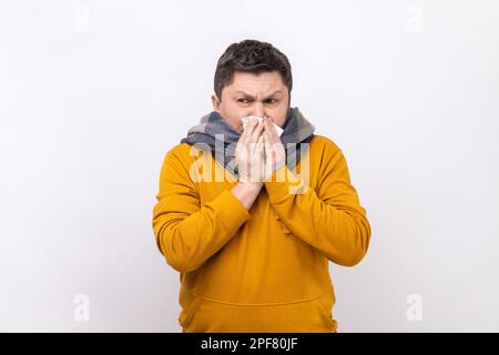 Homme dans le foulard chaud toux éternuant fort dans la serviette, se sentant mal avec le nez qui coule, allergie saisonnière sévère, symptômes de la grippe, port de sweat à capuche de style urbain. Studio d'intérieur isolé sur fond blanc. Banque D'Images