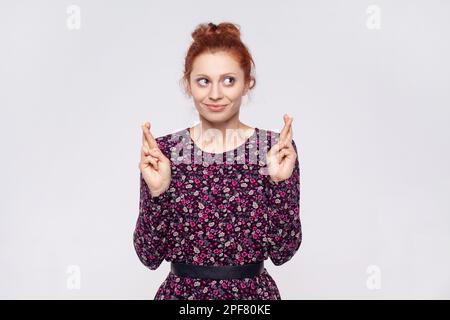 Portrait de la femme à tête rouge portant la robe fait le vœu croit à la bonne chance, les espoirs les rêves deviennent réalité, a l'expression d'espoir, anticipe pour quelque chose. Prise de vue en studio isolée sur fond gris. Banque D'Images