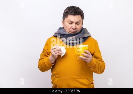 Soins à domicile. Homme malade de la grippe dans un foulard tenant une tasse et un tissu pour le nez qui coule, prenant des médicaments avec du thé chaud, souffrant de fièvre, portant un sweat à capuche de style urbain. Studio d'intérieur isolé sur fond blanc. Banque D'Images