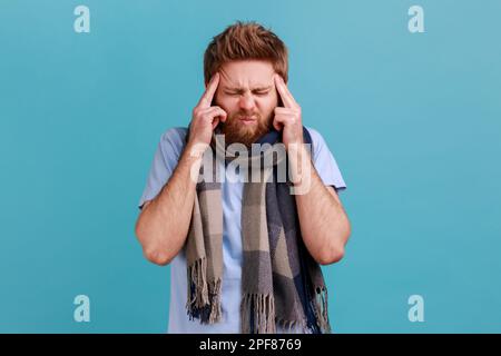 Portrait d'un homme barbu enveloppé d'une écharpe debout avec une grimace douloureuse, touchant la tête, souffrant de migraine, malade de la grippe. Studio d'intérieur isolé sur fond bleu. Banque D'Images