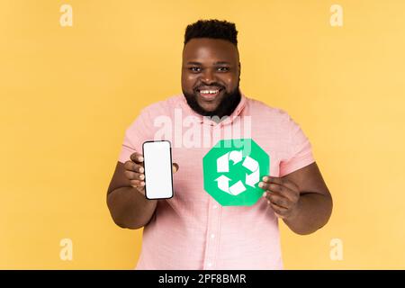 Portrait d'un homme portant une chemise rose montrant un téléphone portable avec écran vierge et symbole de recyclage, être heureux, regardant l'appareil photo avec un sourire crasseux. Studio d'intérieur isolé sur fond jaune. Banque D'Images