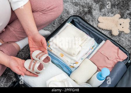Femme enceinte tenant des chaussures pour bébé et emballant le sac de maternité de l'hôpital. Mère pendant la grossesse attendant que bébé prépare une valise de vêtements, de jouets et Banque D'Images