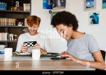 Jeune belle femme africaine et souriant redhead homme tenant et utilisant leur téléphone en train de diring petit déjeuner à la maison Banque D'Images