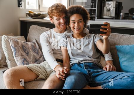 Jeune heureux couple interracial prenant selfie avec téléphone ensemble , tout en tenant les mains et assis sur le canapé dans le salon confortable à la maison Banque D'Images