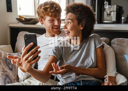 Jeune beau redhead souriant homme embrassant et regardant sa belle amie africaine heureuse avec téléphone, tandis qu'ils sont assis sur un canapé dans la vie confortable Banque D'Images