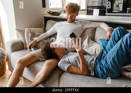 Jeune homme souriant à tête rouge regardant sa belle copine africaine tenant le téléphone et faisant la liaison sur son genou , alors qu'ils parlent sur un canapé Banque D'Images