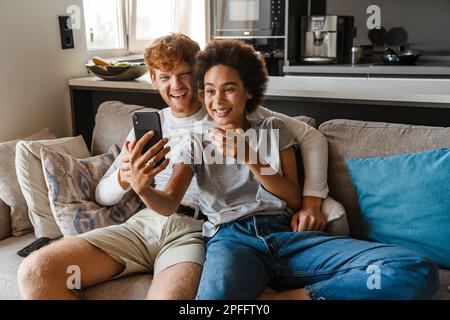 Jeune heureux couple interracial prenant selfie avec téléphone ensemble, tout en étant assis sur un canapé dans le salon confortable à la maison Banque D'Images