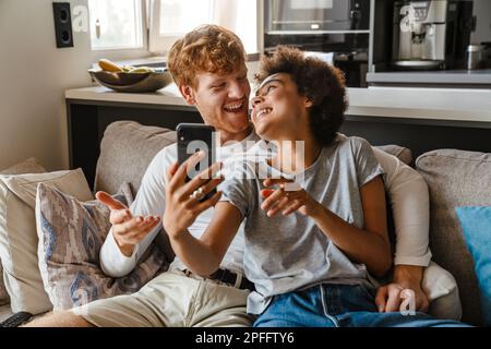 Jeune beau redhead heureux homme embrassant sa belle amie africaine souriante avec téléphone, tandis qu'ils sont assis sur un canapé dans le salon confortable à la maison Banque D'Images