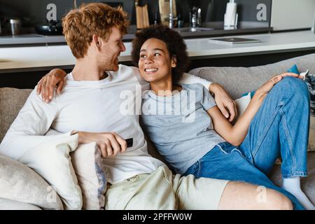 Jeune beau couple heureux assis ensemble sur un canapé à la maison, embrassant et regardant l'un l'autre. Beau redhead homme et beau sourire africain Banque D'Images