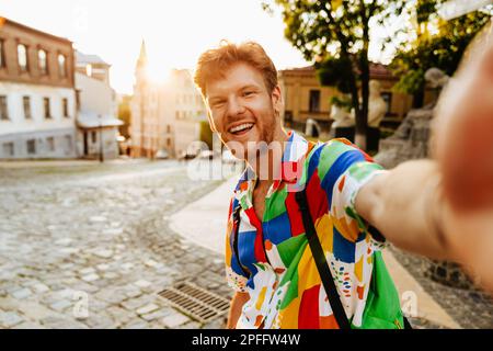 Selfie d'un jeune homme souriant et souriant à tête rouge dans une chemise colorée debout dans la rue Banque D'Images