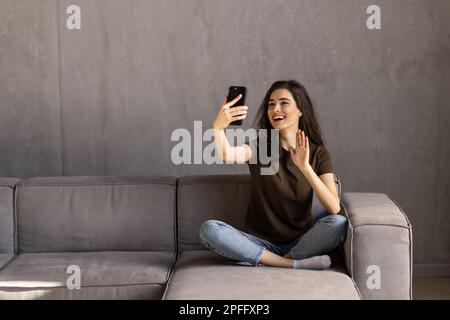 Femme souriante assise sur le canapé de cuisine parlant par videocall datant en ligne regardant le téléphone. Vidéo blogger vlogger enregistrement vlog à la maison. Style de vie vlo Banque D'Images