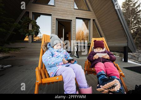 Deux petites filles s'asseyent sur des chaises en terrasse à l'extérieur de la petite maison grillagée dans les montagnes et regardant des dessins animés sur les téléphones mobiles. Banque D'Images