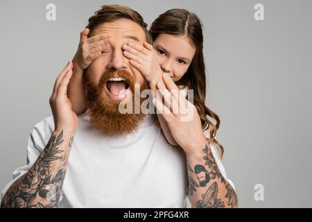 Préteen fille couvrant les yeux de père tatoué avec la bouche ouverte isolée sur gris, image de stock Banque D'Images