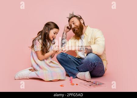 Fille gaie appliquant vernis à ongles sur la main de papa tatoué avec serre-tête couronne sur fond rose, image de stock Banque D'Images