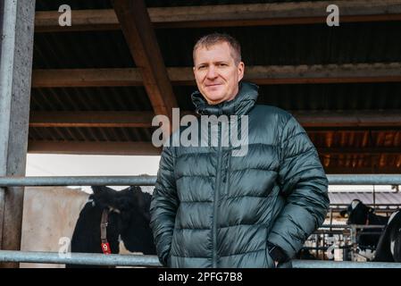 Homme fermier près de ses vaches. Portrait d'un jeune propriétaire de ferme debout dans un long bassin de vache près du corral avec des veaux. Homme dans des vêtements ordinaires vérifie le Banque D'Images