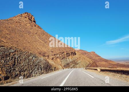 Route asphaltée, petites montagnes de l'Atlas et collines rocheuses des deux côtés, ciel clair au-dessus - paysage typique dans le sud du Maroc Banque D'Images