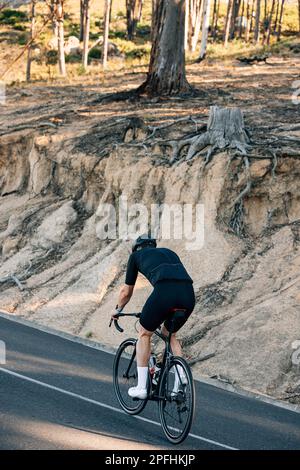 Vue arrière d'un cycliste en tenue de sport noire qui monte sur une colline sur une route de campagne Banque D'Images