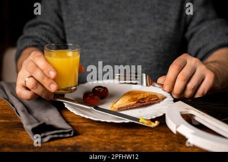 Heure du petit déjeuner. Homme méconnaissable tenant un verre de jus d'orange. Le sandwich se trouve sur l'assiette. Banque D'Images