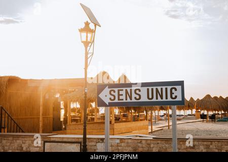 Panneau à sens unique sur la route de bord de mer près de la plage, au lever du soleil. Des parasols en paille peuvent être vus en arrière-plan. Banque D'Images