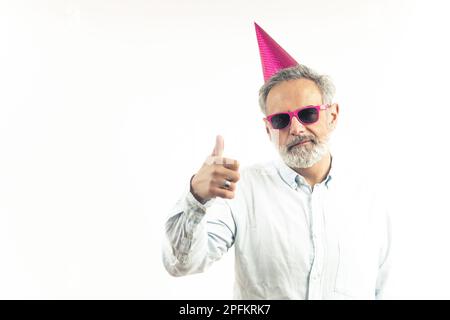 Un homme à barbe aux cheveux gris portant une casquette de fête et des lunettes roses fait un geste de pouce vers le haut. Photo de haute qualité Banque D'Images