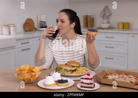 Femme en surpoids buvant du cola et tenant un gâteau dans la cuisine. Aliments malsains Banque D'Images