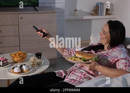 Bonne femme en surpoids avec de la nourriture malsaine regardant la télévision sur le canapé à la maison Banque D'Images