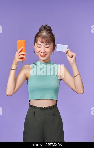 Femme optimiste souriant tout en tenant le smartphone et la carte de crédit isolé sur fond violet Banque D'Images