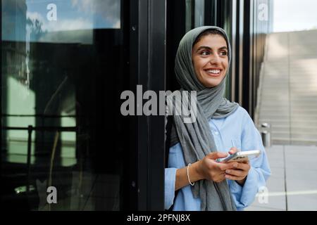 Gaie femme musulmane d'âge moyen portant le hijab au téléphone tout en se tenant debout à l'extérieur Banque D'Images
