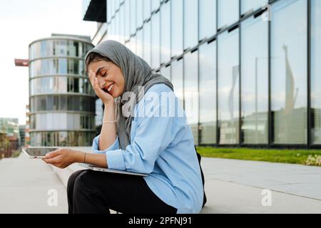 Femme musulmane d'âge moyen souriante portant le hijab tenant un téléphone portable tout en étant assise à l'extérieur dans la rue de la ville Banque D'Images