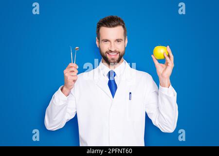 Portrait de dentiste toothy réussi en blouse de laboratoire blanche, cravate bleue, matériel de maintien et pomme verte dans les mains, isolé sur fond gris, recommander Banque D'Images