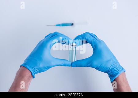 Sur fond blanc isolé, les mains des médecins en gants médicaux bleus représentent le geste de la forme du cœur. Le médecin tient une ampoule avec le vaccin contre v Banque D'Images