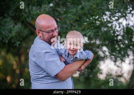 Homme chauve avec des lunettes jette l'enfant dans l'air du ciel. Père en jeans joue, embrassez son fils dans la nature en dehors de la ville. Un petit garçon rit, s'amuse bien Banque D'Images