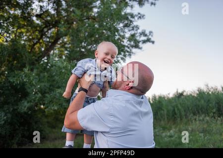 Homme chauve avec des lunettes jette l'enfant dans l'air du ciel. Père en jeans joue, embrassez son fils dans la nature en dehors de la ville. Un petit garçon rit, s'amuse bien Banque D'Images