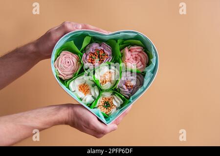 Sur fond isolé orange, les mains mâles tiennent une boîte cadeau en forme de coeur avec des fleurs de guimauve douces. Naturel fait à la main. Dessert sans sucre dans le for Banque D'Images