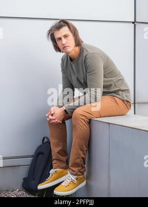 Homme millénaire aux cheveux longs et au look tendu près du mur gris. Un homme sérieux dans des vêtements décontractés aux tons de la terre est à la tête dans l'appareil photo. Banque D'Images