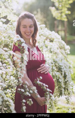 Jeune femme mariée s'attendant bientôt à un bébé, elle est dans le dernier trimestre. Elle est dans le parc, entre les fleurs. Banque D'Images