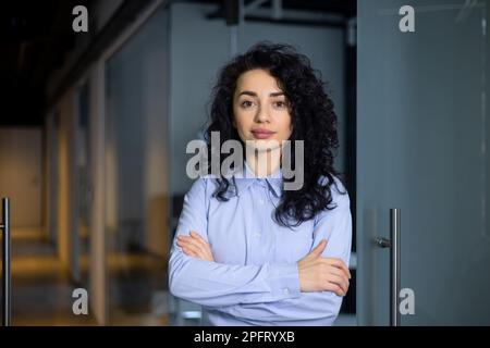 Portrait d'une femme sérieuse patron à l'intérieur du bureau de la société d'affaires, femme d'affaires a croisé les bras regardant concentré à l'appareil photo, portant une chemise, une femme hispanique satisfaite et réussie avec des cheveux bouclés. Banque D'Images