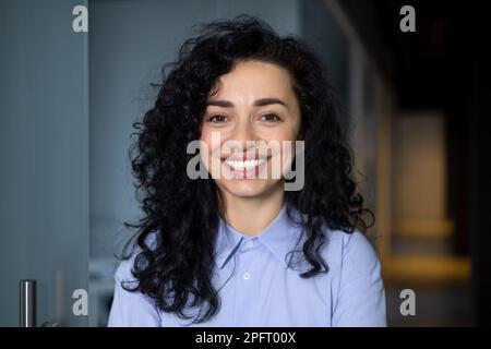 Portrait en gros plan d'une femme d'affaires adulte mature, d'un patron souriant et regardant l'appareil photo, d'une femme hispanique ravie avec des cheveux bouclés dans une chemise bleue à l'intérieur du bureau dans le couloir du hall. Banque D'Images