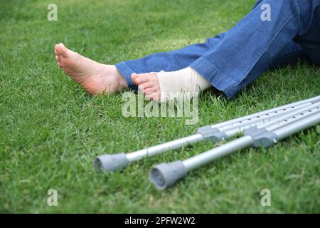 Femme blessée méconnaissable assise sur l'herbe, détail de ses pieds, l'un d'eux avec un bandage, et une paire de béquilles à son côté. Concept : recove Banque D'Images