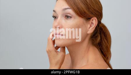 Portrait d'une femme d'âge moyen avec cheveux rougeâtres tenant la main sur la joue regardant l'appareil photo sur fond gris Banque D'Images