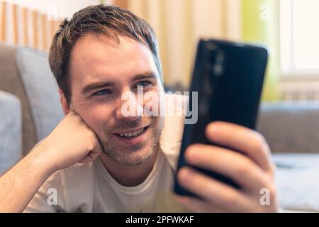 Heureux homme utilisant un smartphone à la maison, il discutant de la messagerie en ligne sur mobile. Un jeune homme se repose sur le canapé et lit les informations d'un smartp Banque D'Images