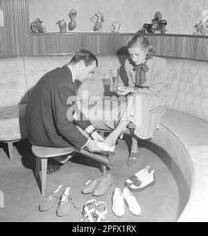 Une jeune femme est bien prise en charge par un commis dans un magasin de chaussures. Le répartiteur essaie différentes chaussures sur les pieds de la dame. Elle a essayé plusieurs paires différentes de chaussures d'été et, à en juger par la photo, elle n'a pas encore trouvé une paire qu'elle pense convenir. Suède 1949 Kristoffersson réf. AS15-1 Banque D'Images