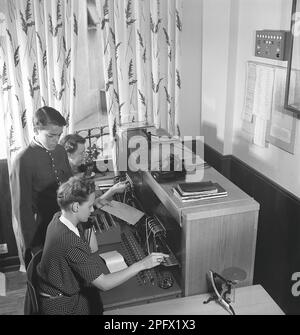 Standard dans les jours anciens. Deux femmes travaillent au standard de l'entreprise. Ils connectent les appels entrants et sortants à la bonne personne à l'aide de cordons reliés aux téléphones respectifs dans un commutateur de cordon. À côté d'elle se tient le garçon de course de la compagnie, soigneusement vêtu d'uniforme et tenant une enveloppe importante. Suède 1944 Kristoffersson réf. K66-4 Banque D'Images